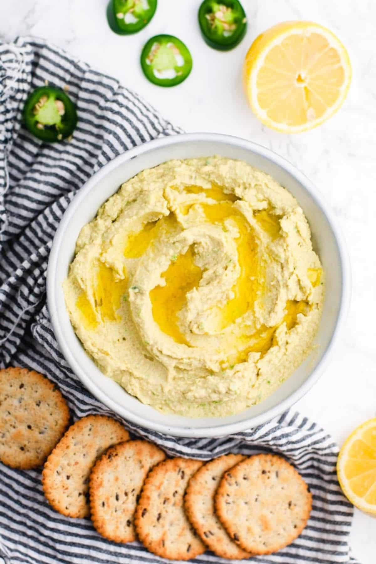 Bowl filled with creamy appearing hummus next to crackers and sliced jalapenos.