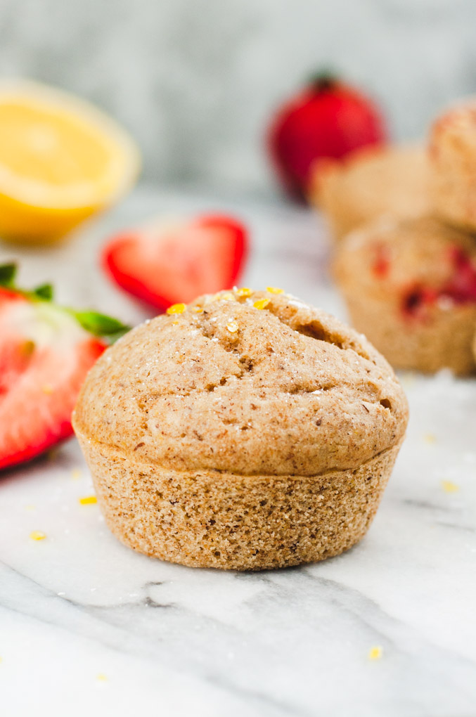 A single strawberry muffin in the foreground with more blurred muffins and strawberries in the background