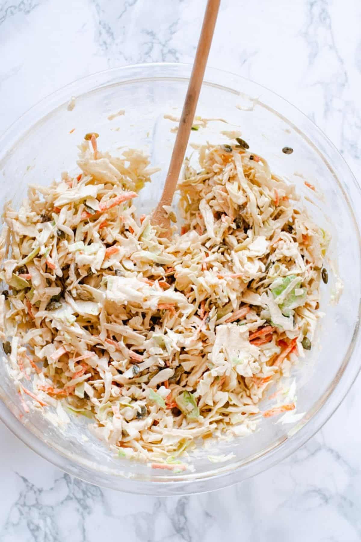 Tahini coleslaw in a large glass bowl on a marble background. A wooden spoon is resting in the bowl.