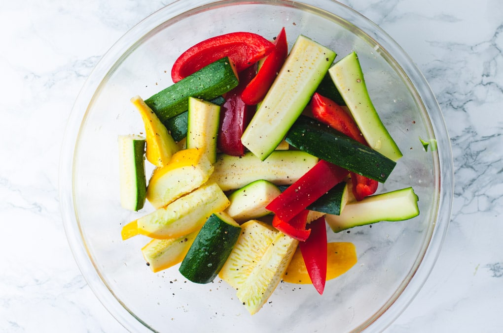 Sliced uncooked zucchini, squash, and red peppers in a glass mixing bowl. 