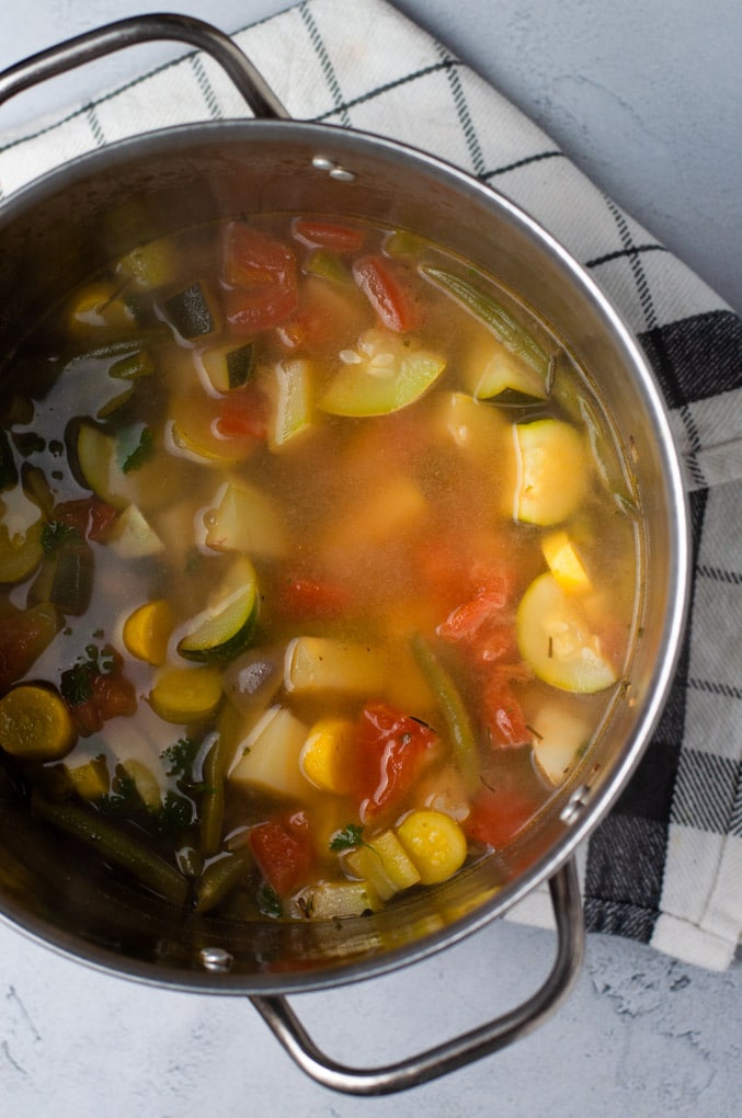 Vegetable soup in metal stockpot on a white and black striped cloth and gray background.