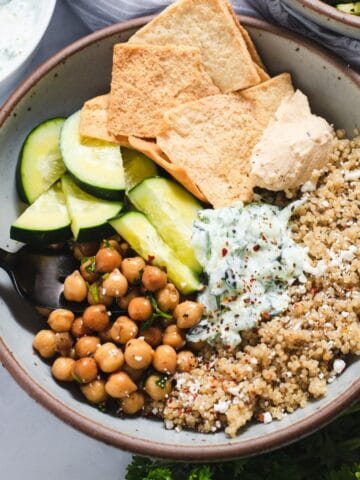 Gray speckled ceramic bowl showing separated quinoa, chickpeas, pita chips, cucumber and tzatziki sauce.
