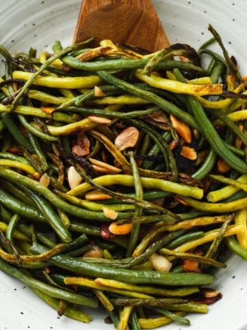 One recipe of green beans almondine in a white ceramic bowl with a wooden serving spoon.