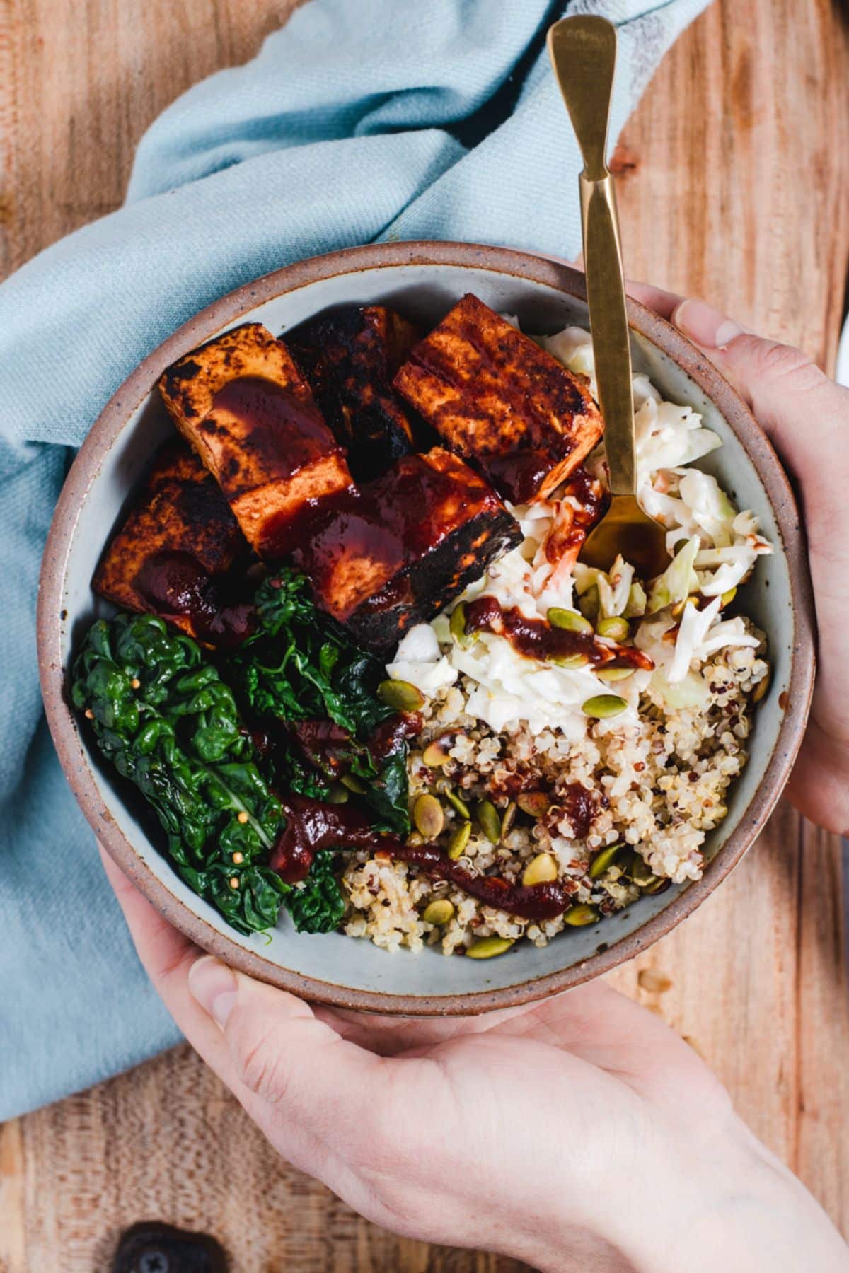 Hands wrapped around a blue bowl filled with tofu, kale, quinoa and coleslaw.