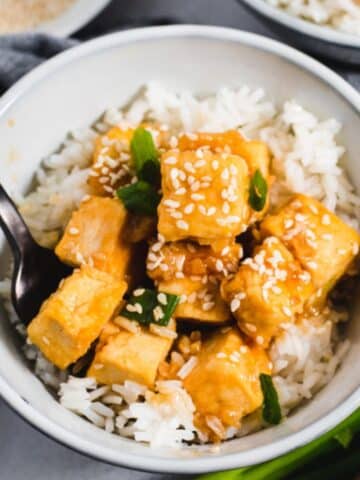 Sesame tofu on white rice in a gray bowl with green onions in the background.