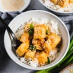 Sesame tofu on white rice in a gray bowl with green onions in the background.