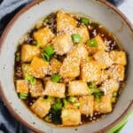 Gray bowl filled with sesame tofu, sliced green onions, and sesame sauce.