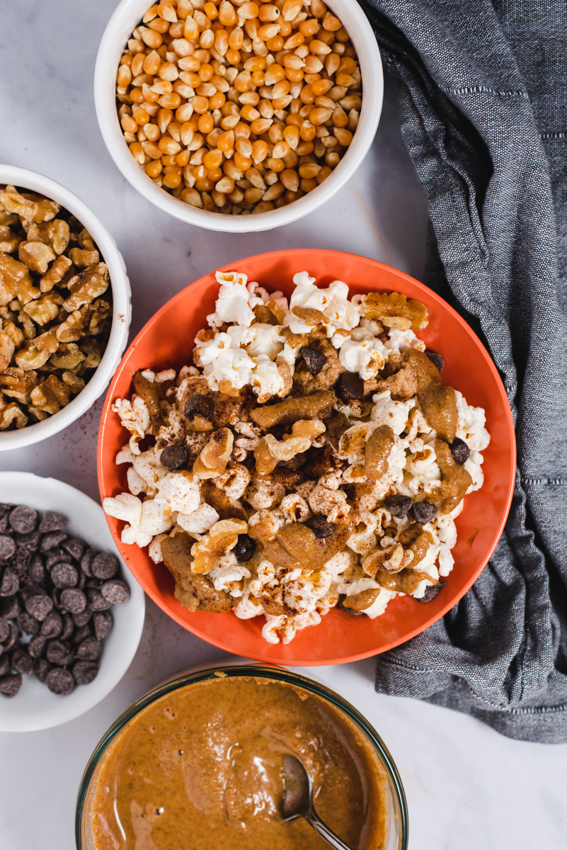 Orange popcorn filled bowl next to bowls filled with chocolate chips, popcorn kernels and walnuts.