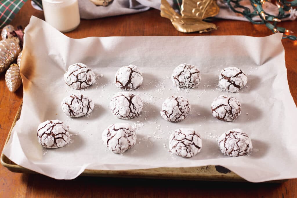 Twelve baked chocolate crinkles lined up on a parchment paper lined baking sheet.