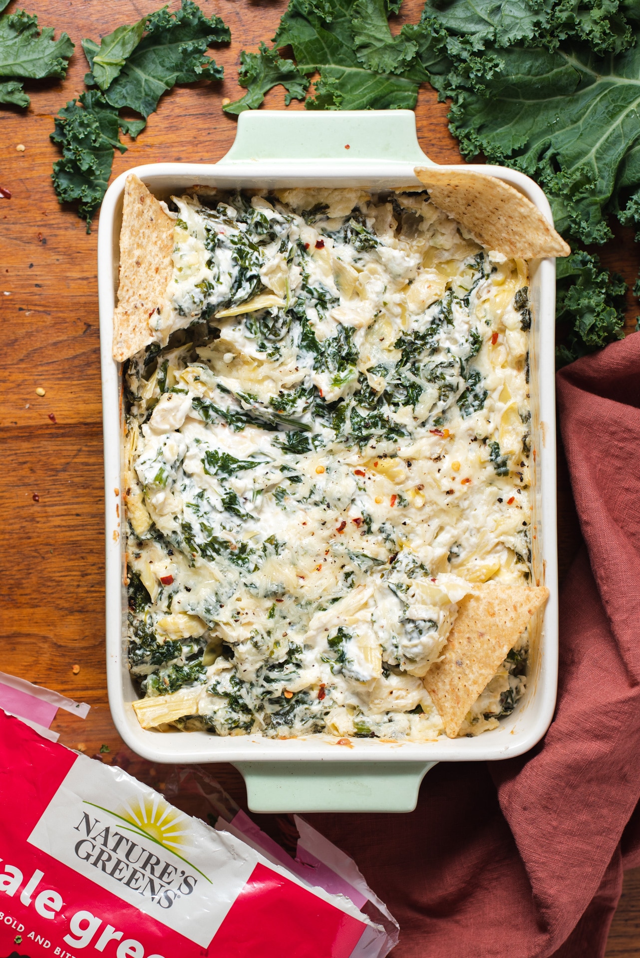Kale artichoke dip in rectangle casserole dish with bag of Nature's Greens in bottom left corner.