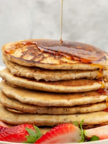 Syrup being poured onto a stack of pancakes surrounded by fresh strawberries.
