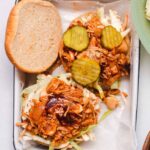 Two open faced sandwiches topped wiht coleslaw, jackfruit and pickles on a white tray