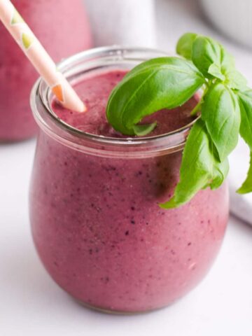 Small jar filled with purple smoothie, a sprig of basil and a paper straw
