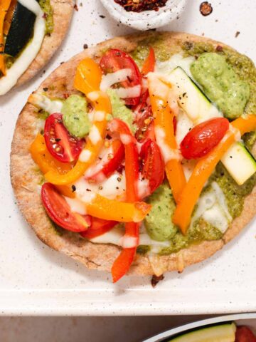 Pita pizza with peppers and zucchini on a white sheet pan next to bowl of red pepper flakes