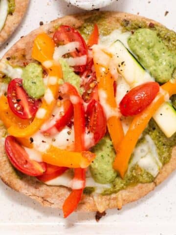 Pita bread pizza with peppers and zucchini on a white sheet pan next to bowl of red pepper flakes.
