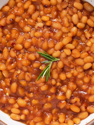 Close up of orange-brown baked beans with a sprig of rosemary on top in a white bowl