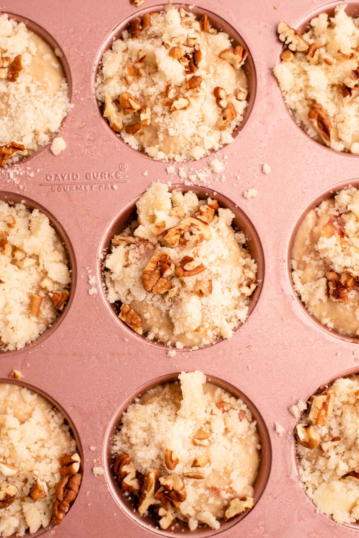 Unbaked muffins topped with crumb topping and pecans in pink muffin tin