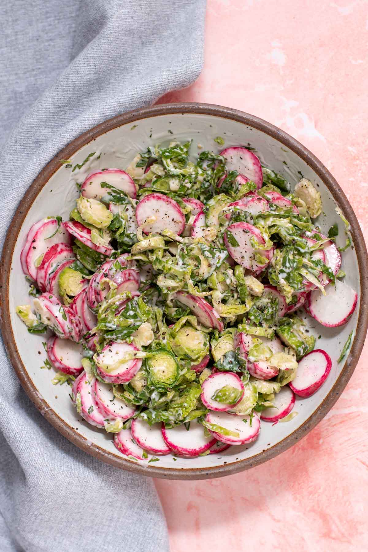 Sliced radishes and brussel sprout slaw in a white speckled bowl on a pink background