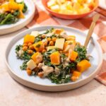 Kale and butternut squash salad on a white plate with bowl of sliced apple in the background.