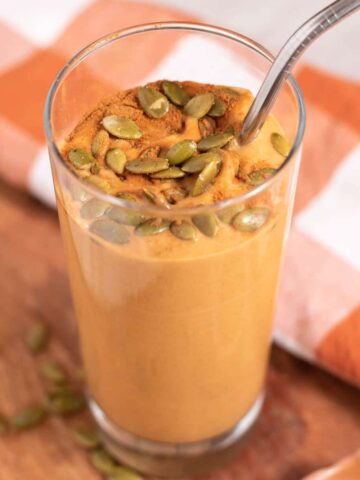 Tall glass of orange smoothie topped with pumpkin seeds and cinnamon on a wood and orange checked background