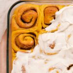 Glass pan of frosted pumpkin cinnamon rolls with two unfrosted cinnamon rolls at the top.