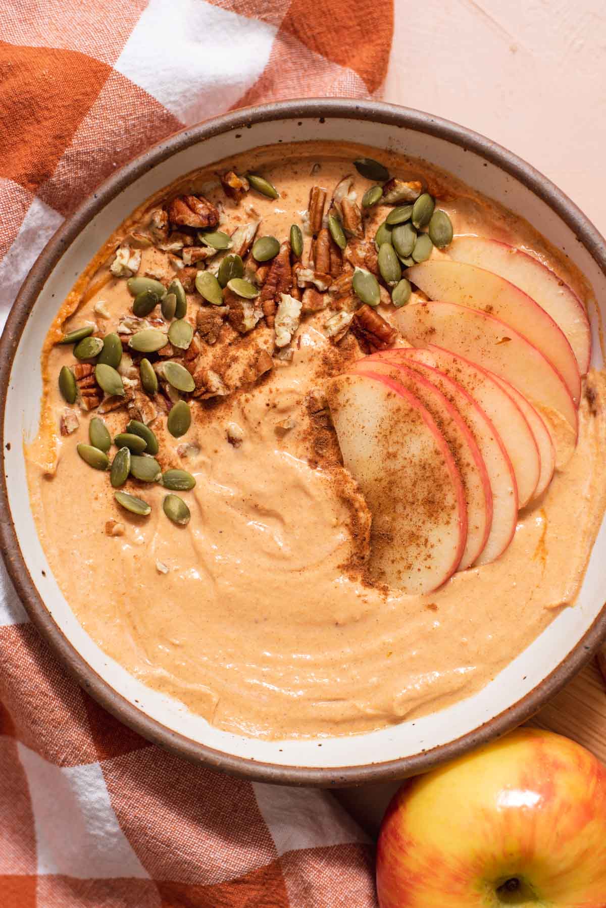 Overhead view of creamy orange dip in a white bowl with pumpkin seeds and apple slices