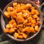 Gray bowl filled with roasted butternut squash pieces next to a green cloth.