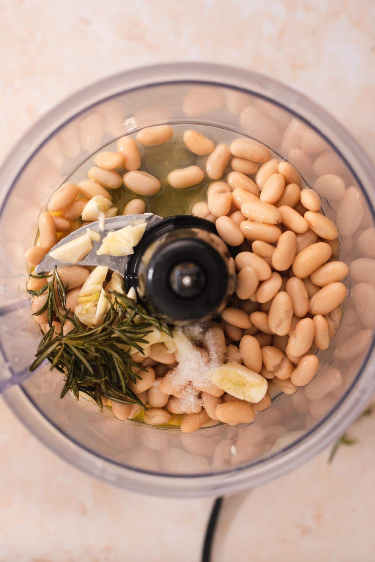 Overhead view of food processor filled with white beans, rosemary and garlic
