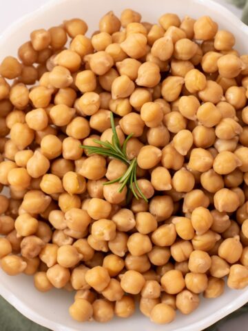 Close up of chickpeas in a white bowl with a sprig of rosemary.