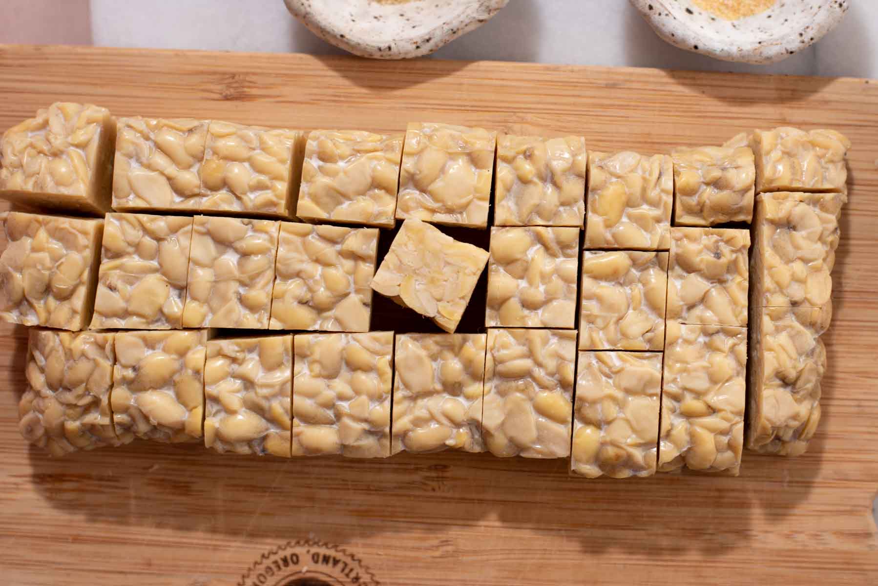 Block of tempeh cut into slices on a wood cutting board.