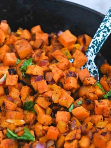 Blue speckled serving spoon lifting up a portion of sweet potato hash from a cast iron skillet.