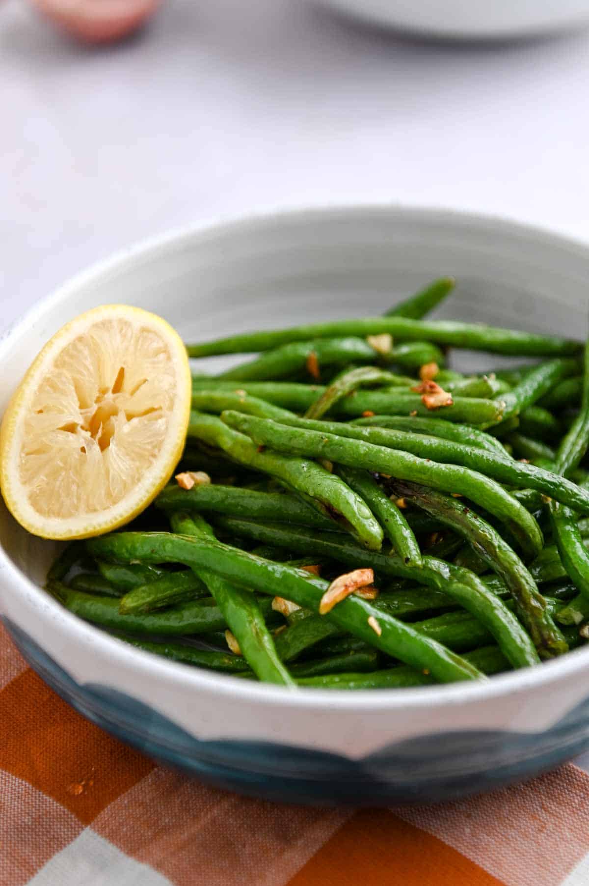 Bowl of green beans with roasted garlic and half a lemon resting on the green beans.
