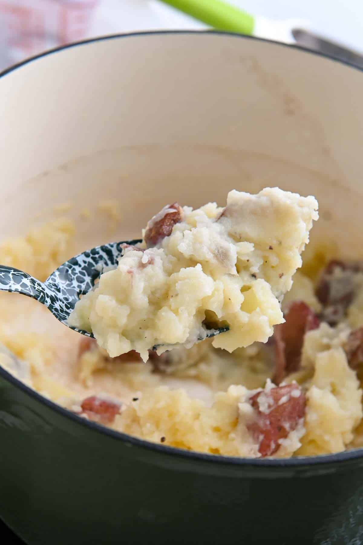 Blue specked metal spoon lifting a serving of mashed potatoes from a Dutch oven.