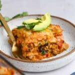 Slice of enchilada casserole topped with avocado and cilantro on a white speckled plate.