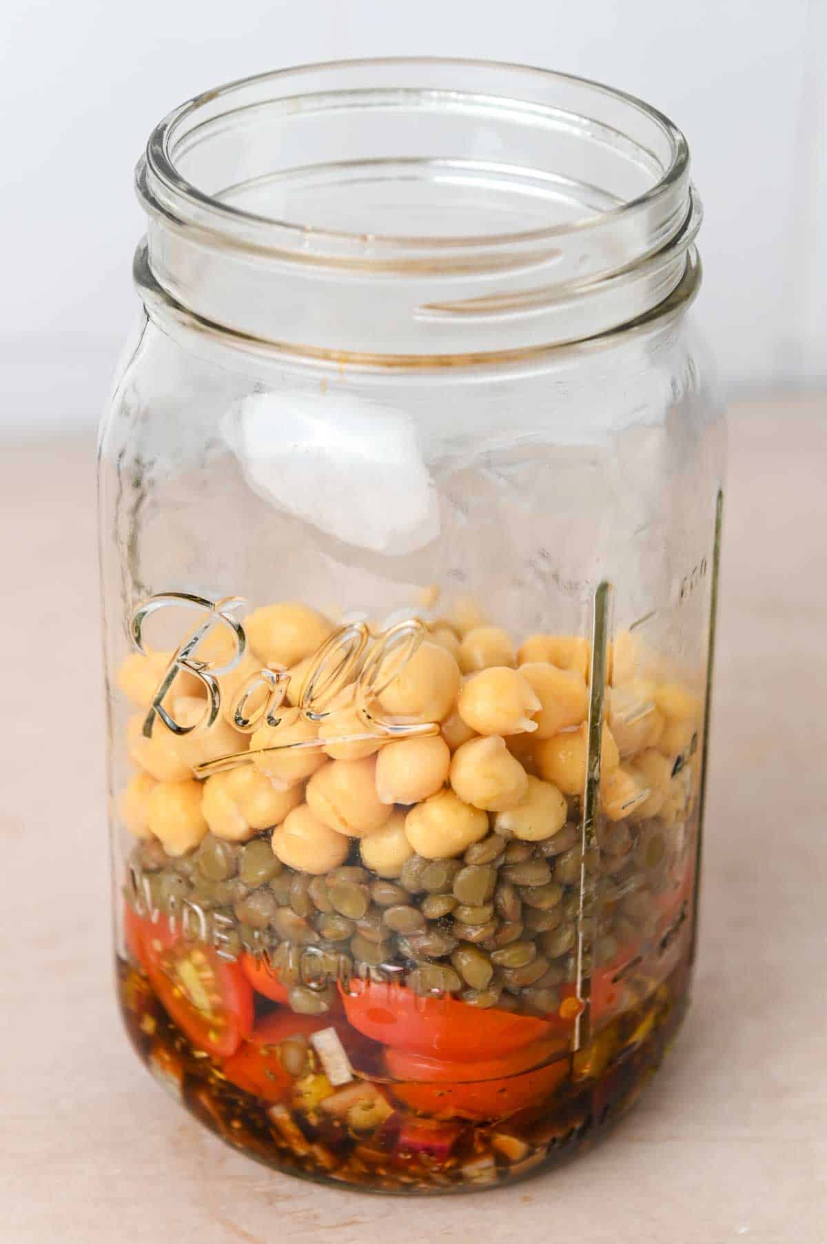 Glass mason jar filled with vinaigrette, tomatoes, onions, lentils, and chickpeas.