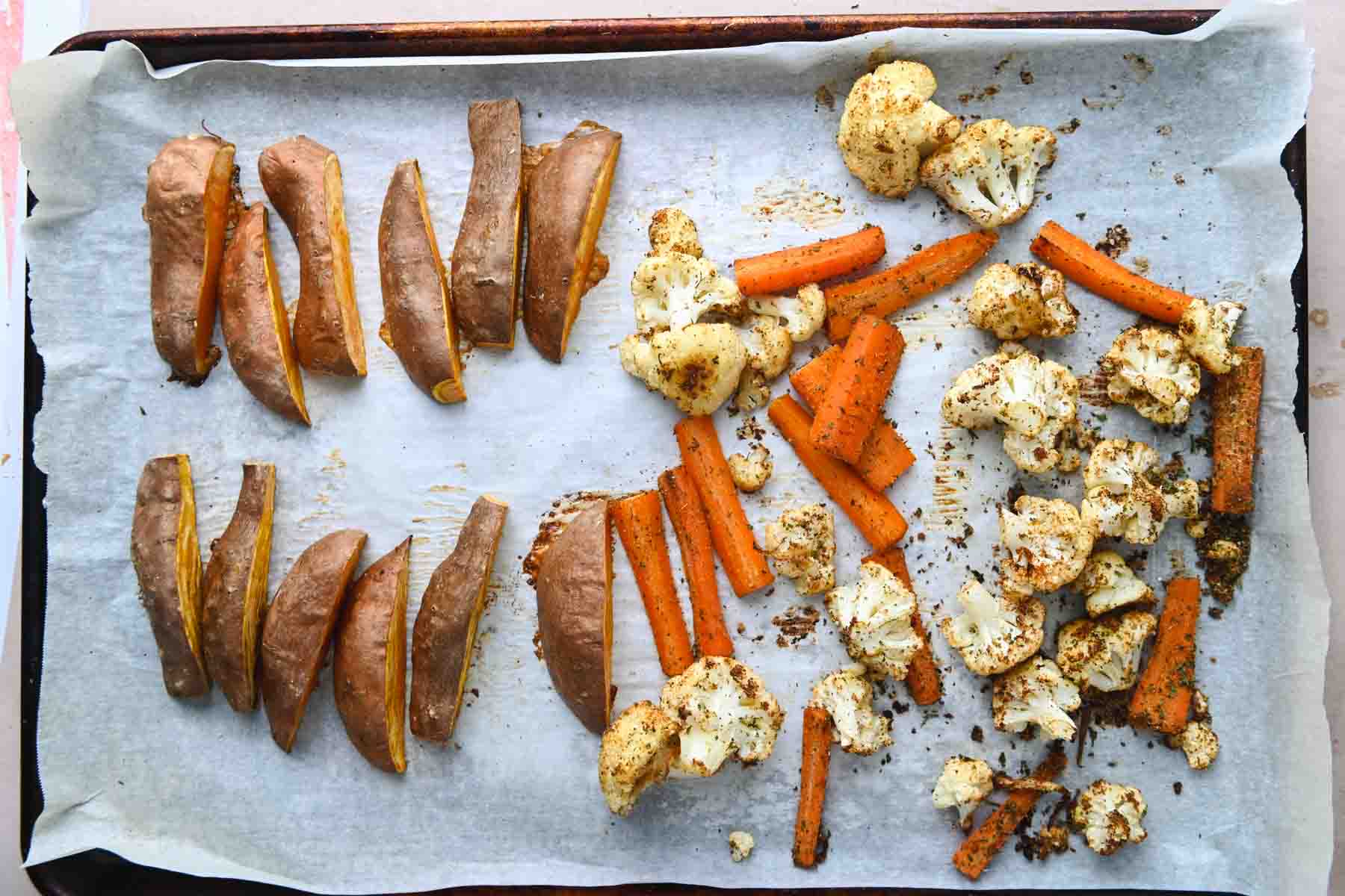 Wedges of sweet potatoes, cauliflower, and carrots on a parchment paper lined sheet pan.
