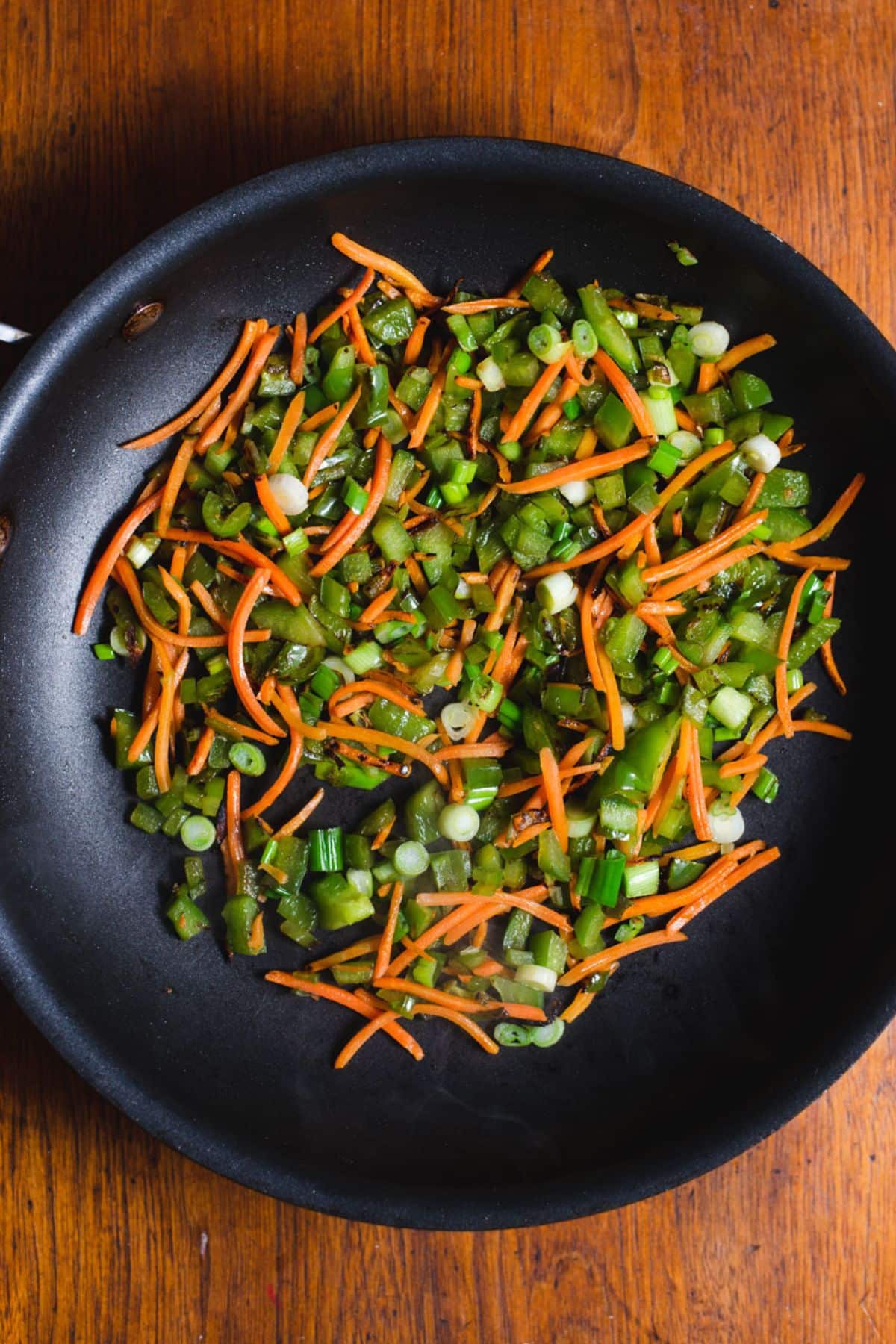 Matchstick carrots, green bell pepper, and green onions cooking in a black skillet.