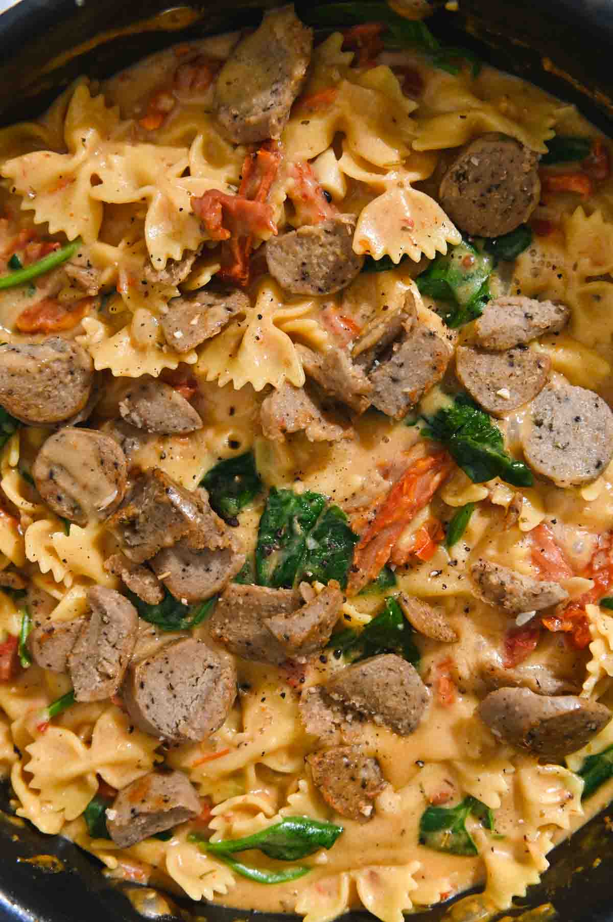 Close up of bowtie pasta with spinach, sun-dried tomato sauce, and sliced Italian sausage.