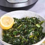 White bowl filled with stewed kale and half of a lemon, with the Instant Pot out of focus in the background.