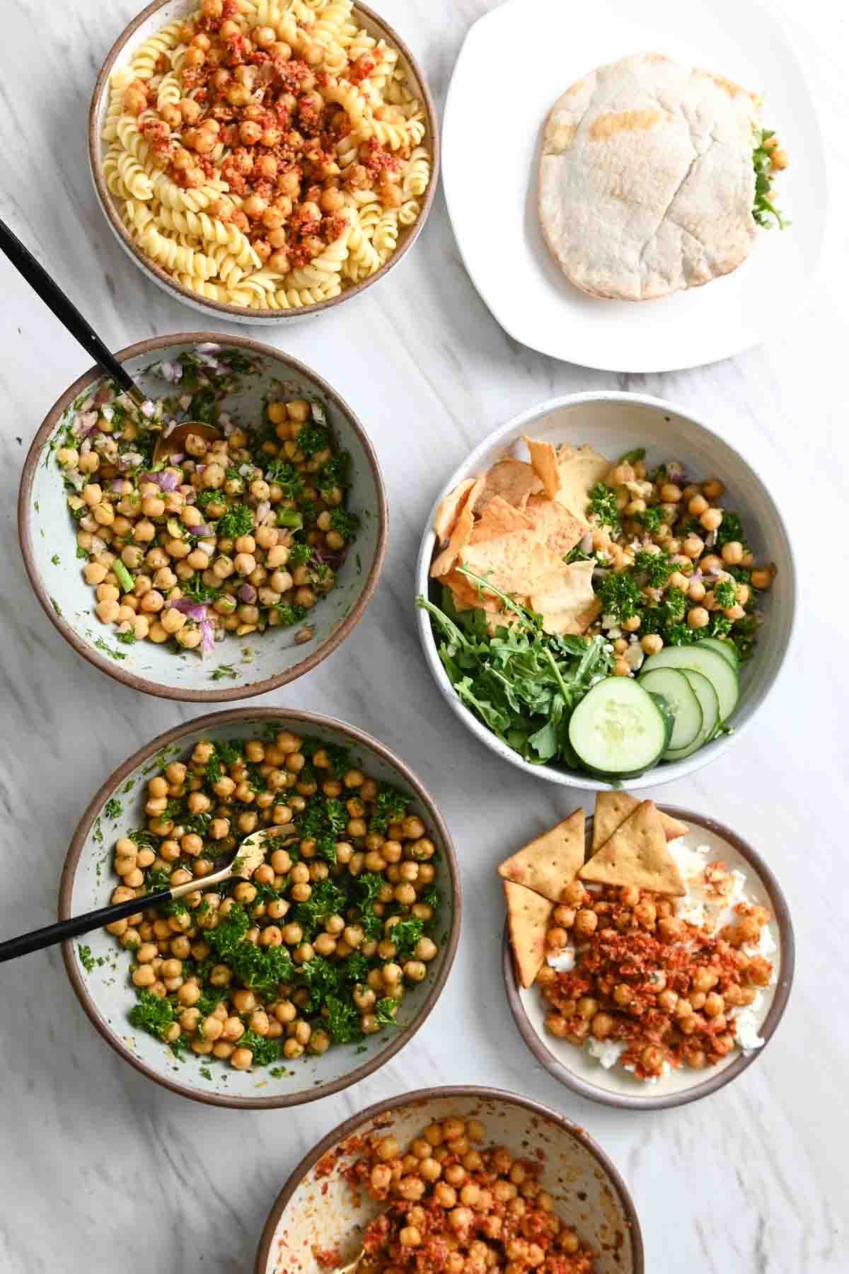 Six bowls and one plate filled with chickpeas, salads, and wraps.