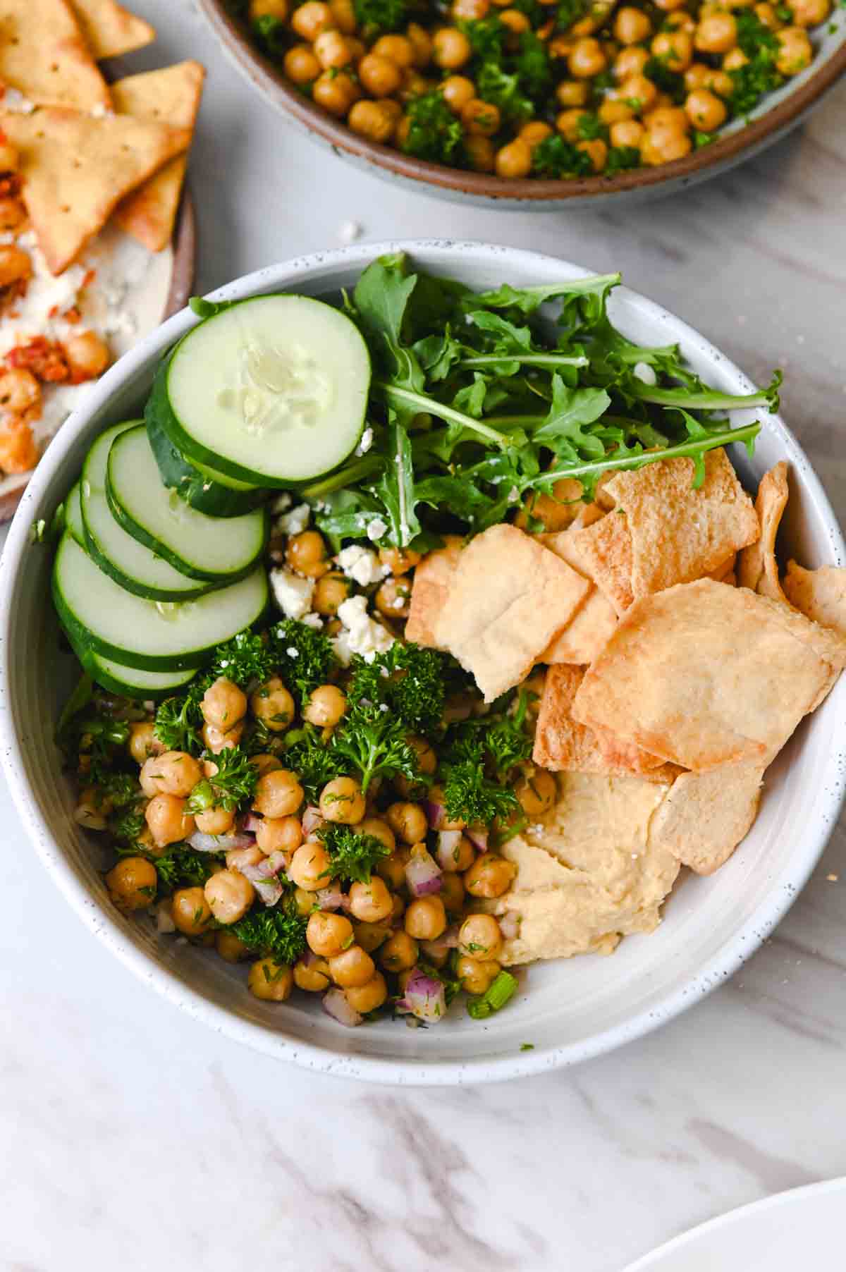 White bowl filled with marinated chickpeas, pita chips, feta, cucumber, and arugula.