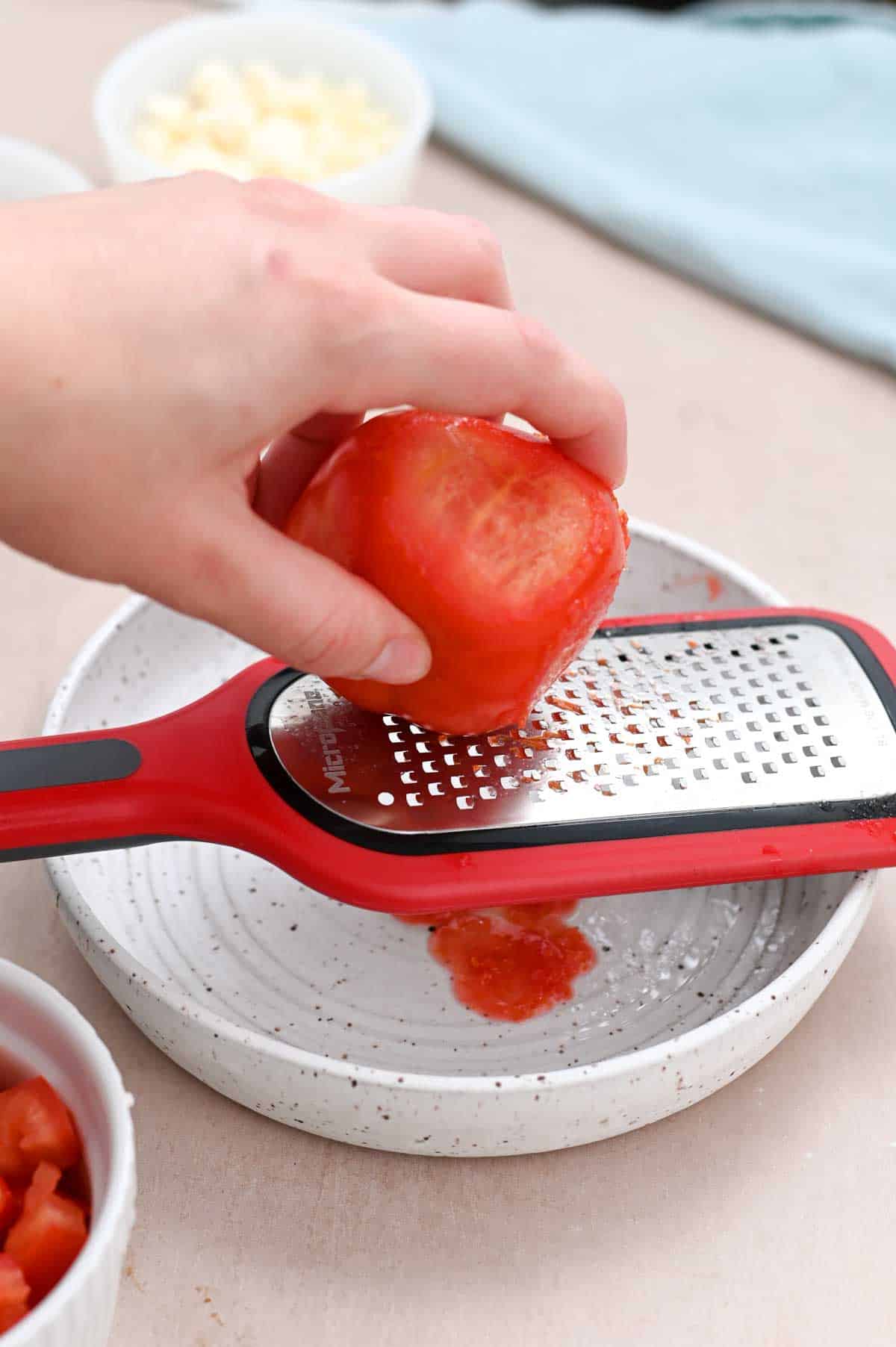 Hand grating a large tomato on a red grater with small holes.