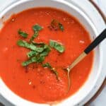 Close up of white bowl filled with vivid red tomato soup and sliced basil.