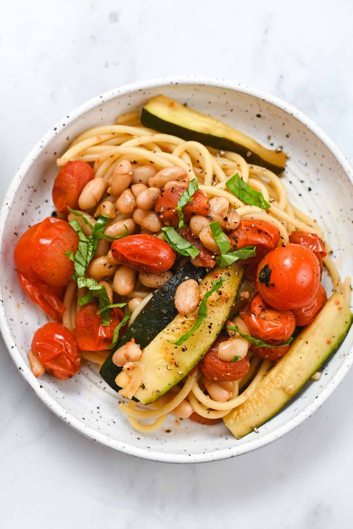 White speckled bowl filled with cherry tomato, zucchini, white beans, and pasta.