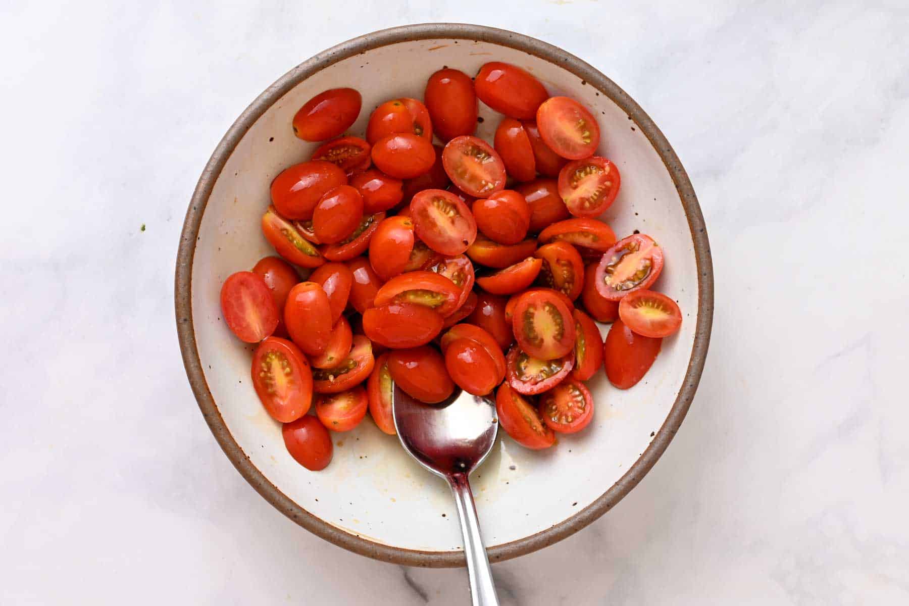 Brown-rimmed white bowl filled with cherry tomatoes tossed in balsamic vinegar and a metal spoon.