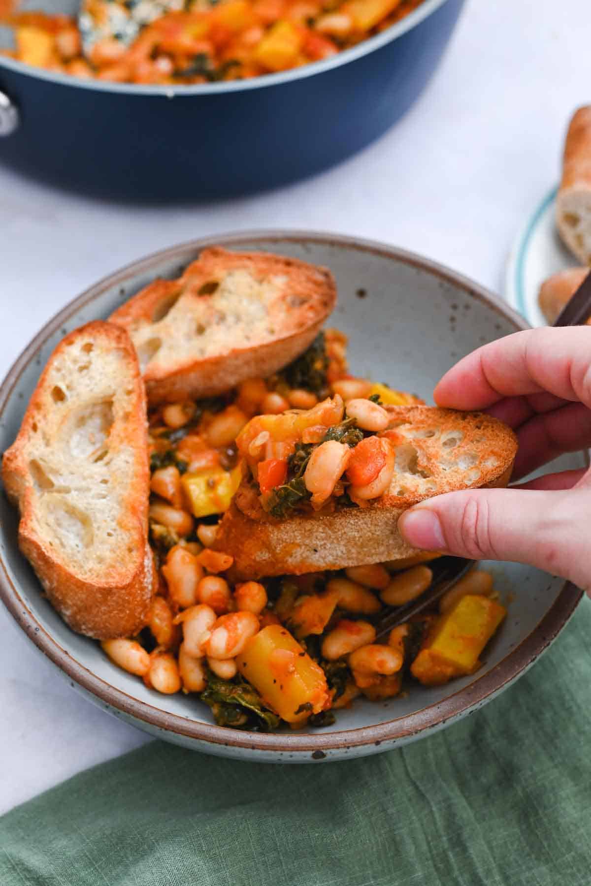Hand lifting a piece of toast with beans on top out of a bowl of white beans.