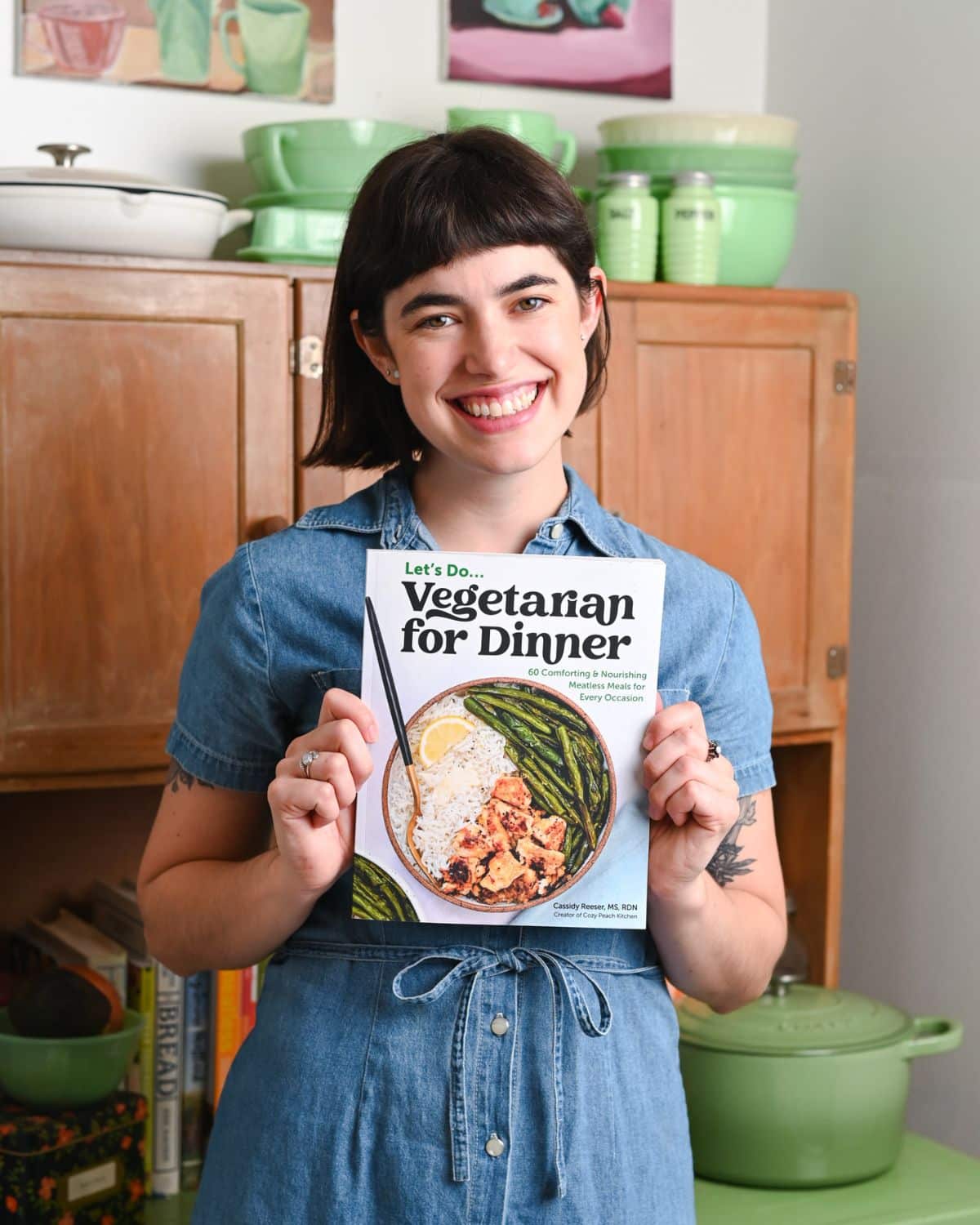Woman with brown hair in a denim dress holding a cookbook and smiling.