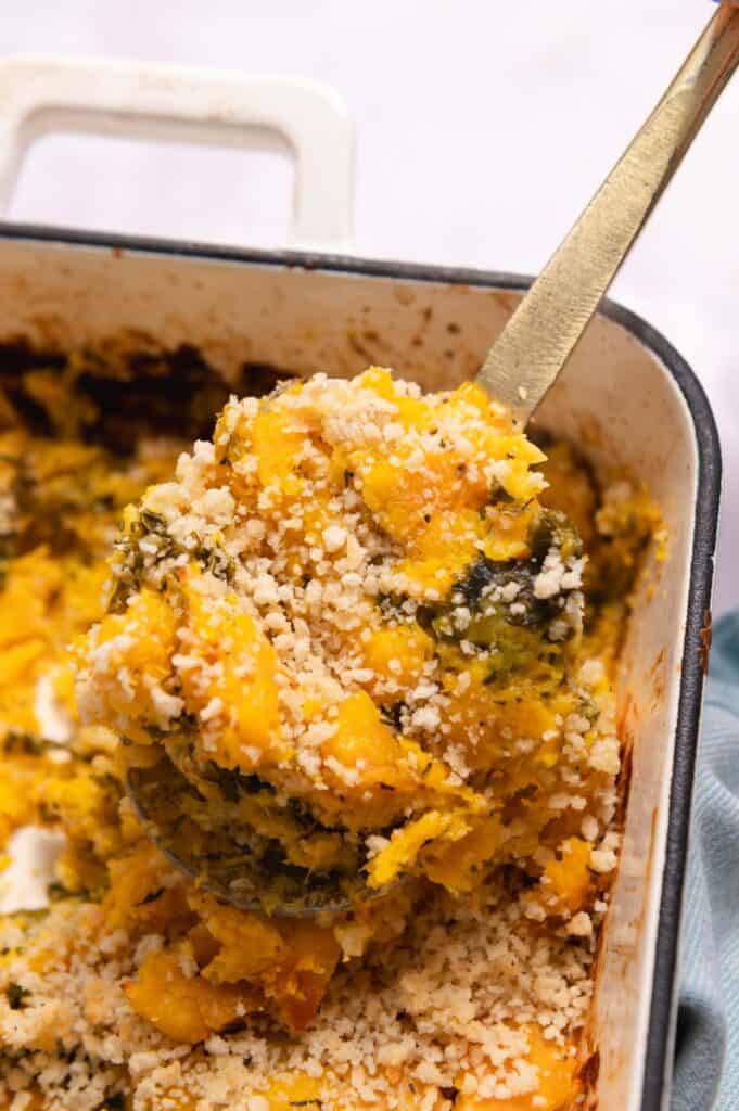 Gold serving spoon lifting a portion of butternut squash casserole out of the baking dish.