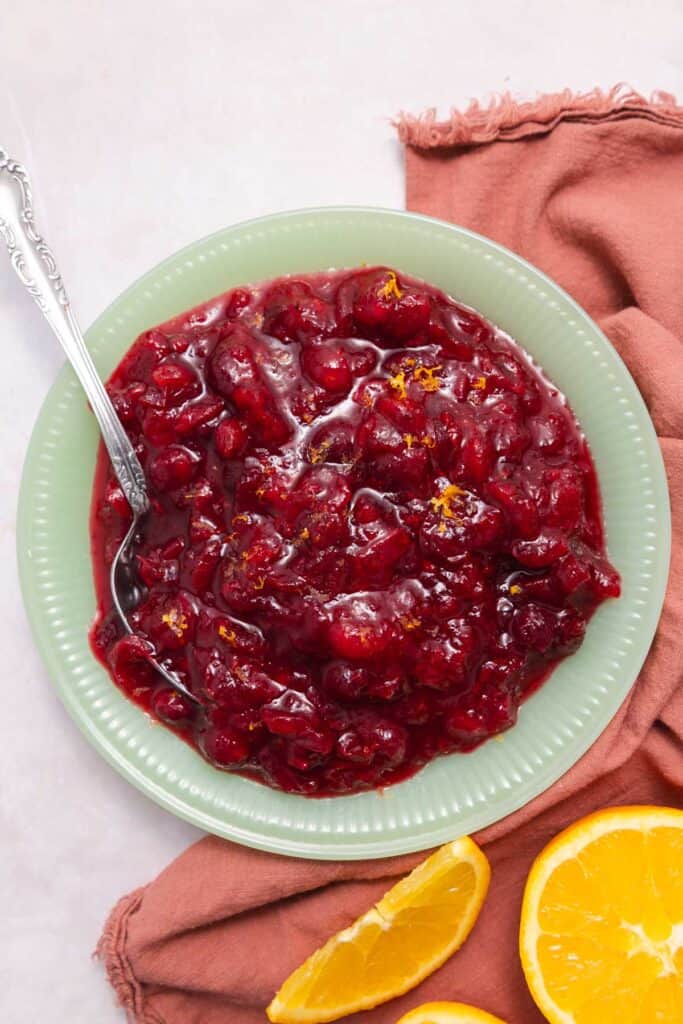 Jade green bowl filled with cranberry sauce next to slices of orange.