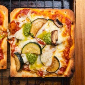 Close up of a square of flatbread pizza with sliced zucchini and dollops of pesto on it.